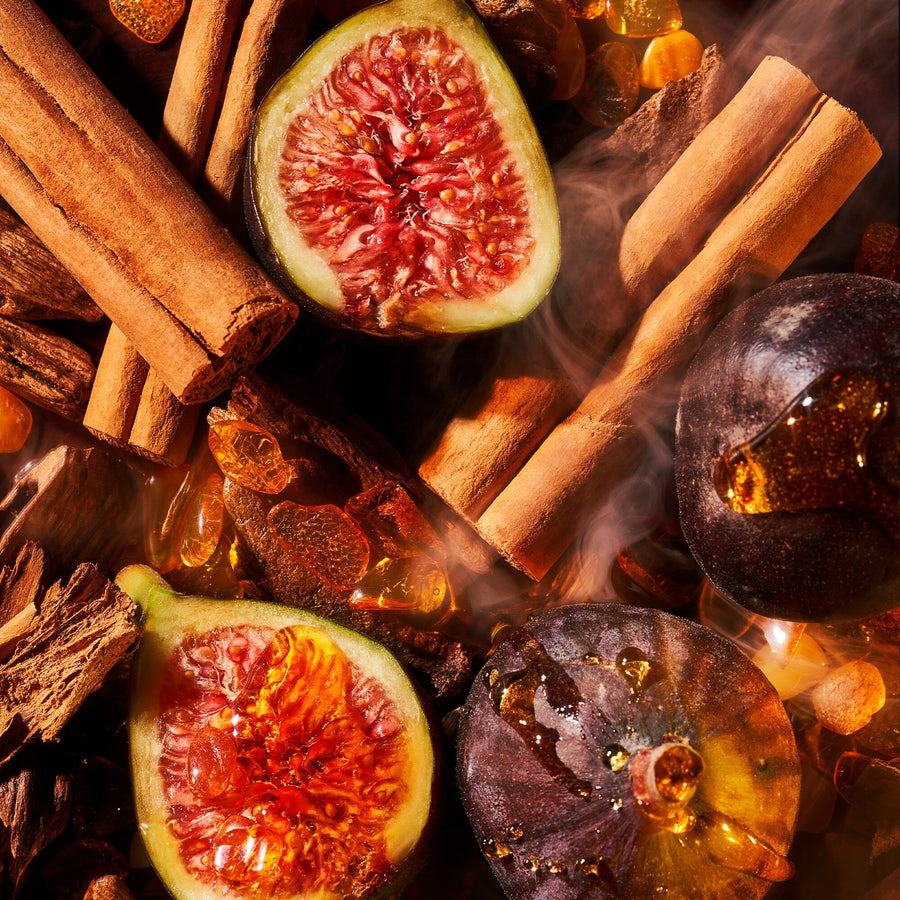 A close-up of halved figs, cinnamon sticks, and an Amber Aoud candle by Roja Parfums creates a warm scene. Smoke wisps add to the iconic scent, while rich figs and textured cinnamon enhance its warmth.