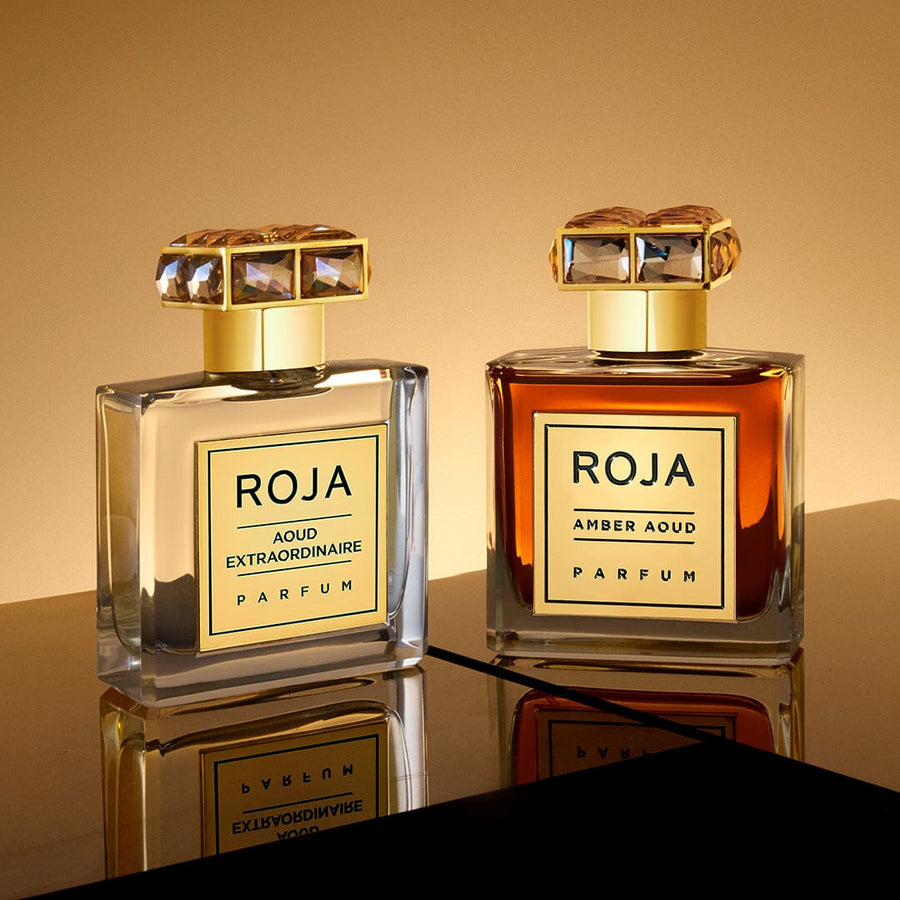 Two elegant perfume bottles from Roja Parfums are displayed on a reflective surface. The clear bottle reads Aoud Extraordinaire, and the amber one displays Amber Aoud, both part of The Aoud Layering Gift Set, featuring decorative square caps against a warm, golden background.