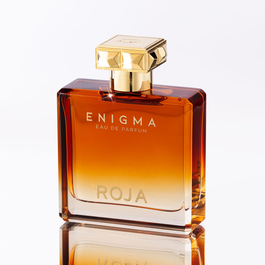 A glass bottle of Enigma Pour Homme by Roja Parfums features a gradient amber color, gold cap, and reflective base. The words ENIGMA and ROJA are printed on the front. It exudes tobacco warmth with a vanilla hint, evoking sophistication similar to fine cognac.