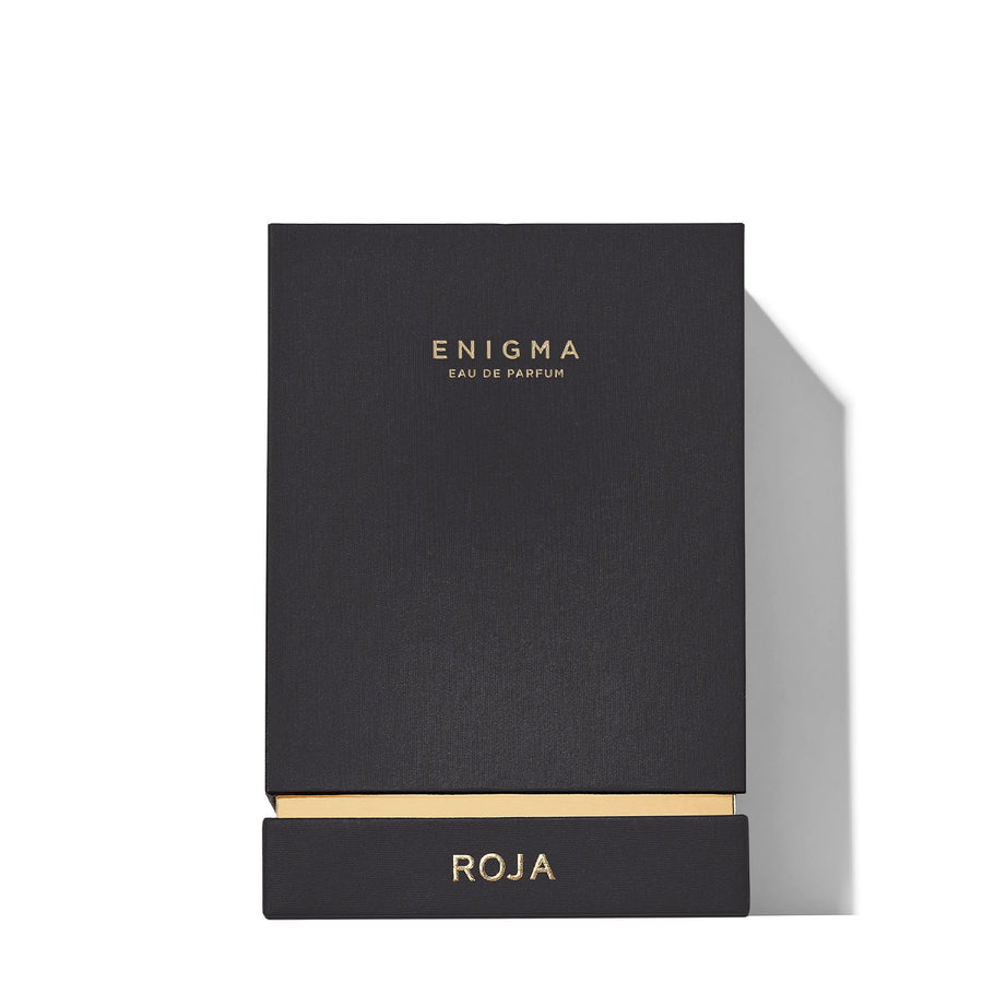 A black fragrance box, marked with ENIGMA POUR HOMME in gold, features the name ROJA PARFUMS on its elegant pull-out drawer. Its sleek design and hints of cognac warmth stand out against a crisp white background.