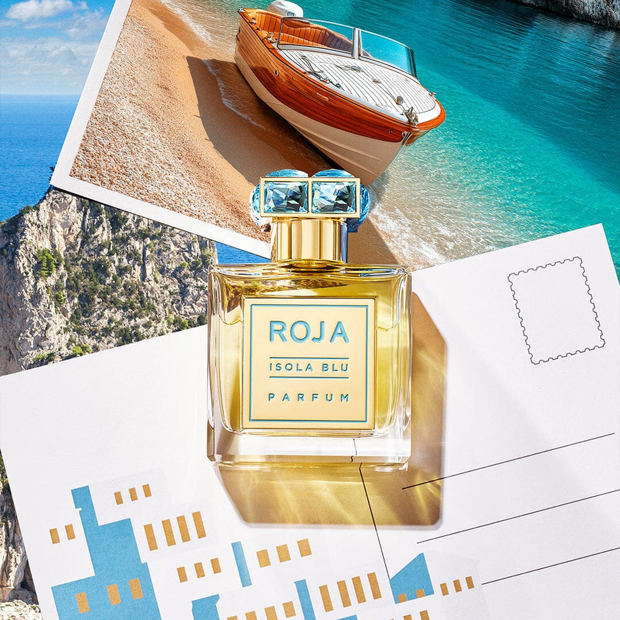 A bottle of Isola Blu by Roja Parfums, a citrus scent, rests on a scenic beach and boat postcard. Its shadow blends with the backdrop, reminiscent of a luxury vacation with cliffside and ocean views.