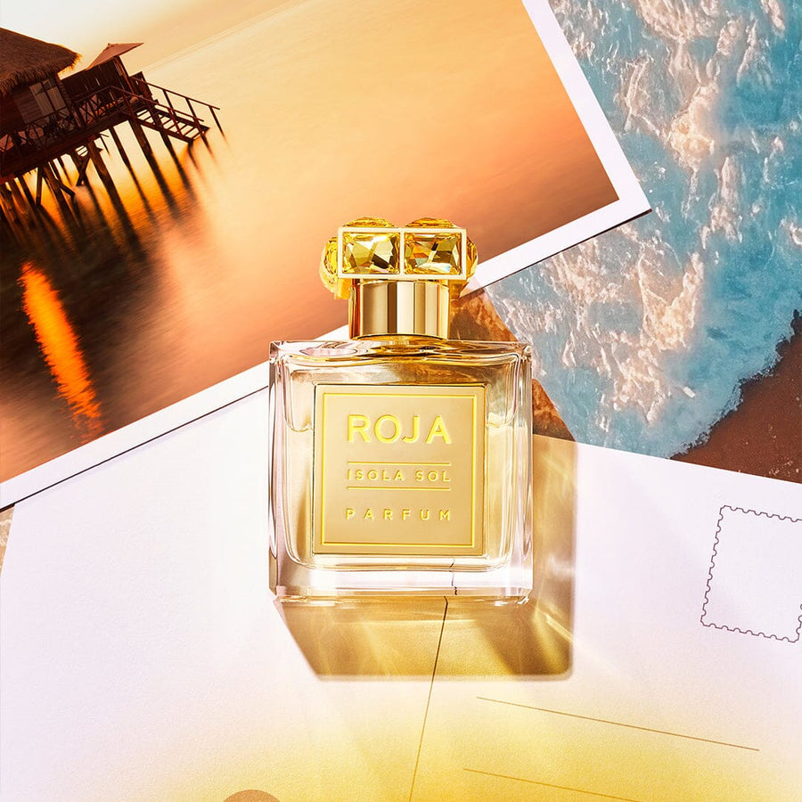 A bottle of Isola Sol by Roja Parfums sits on two postcards, evoking island vibes; one shows a stunning sunset overwater bungalows, and the other, a clear blue ocean with gentle waves. This Roja Dove creation is warm and inviting.